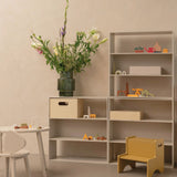 A modern modular shelving unit in a warm beige tone, styled with a large green vase of flowers, wooden storage boxes, colorful children's toys, and a small yellow step stool in a minimalist room.