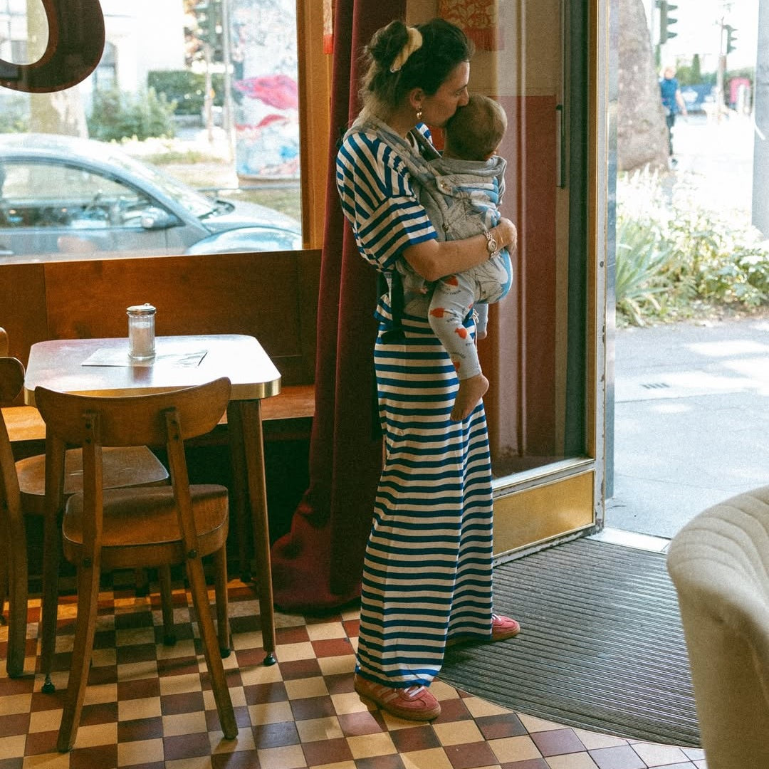 Woman in a striped dress holding a baby in a sling inside a cafe.