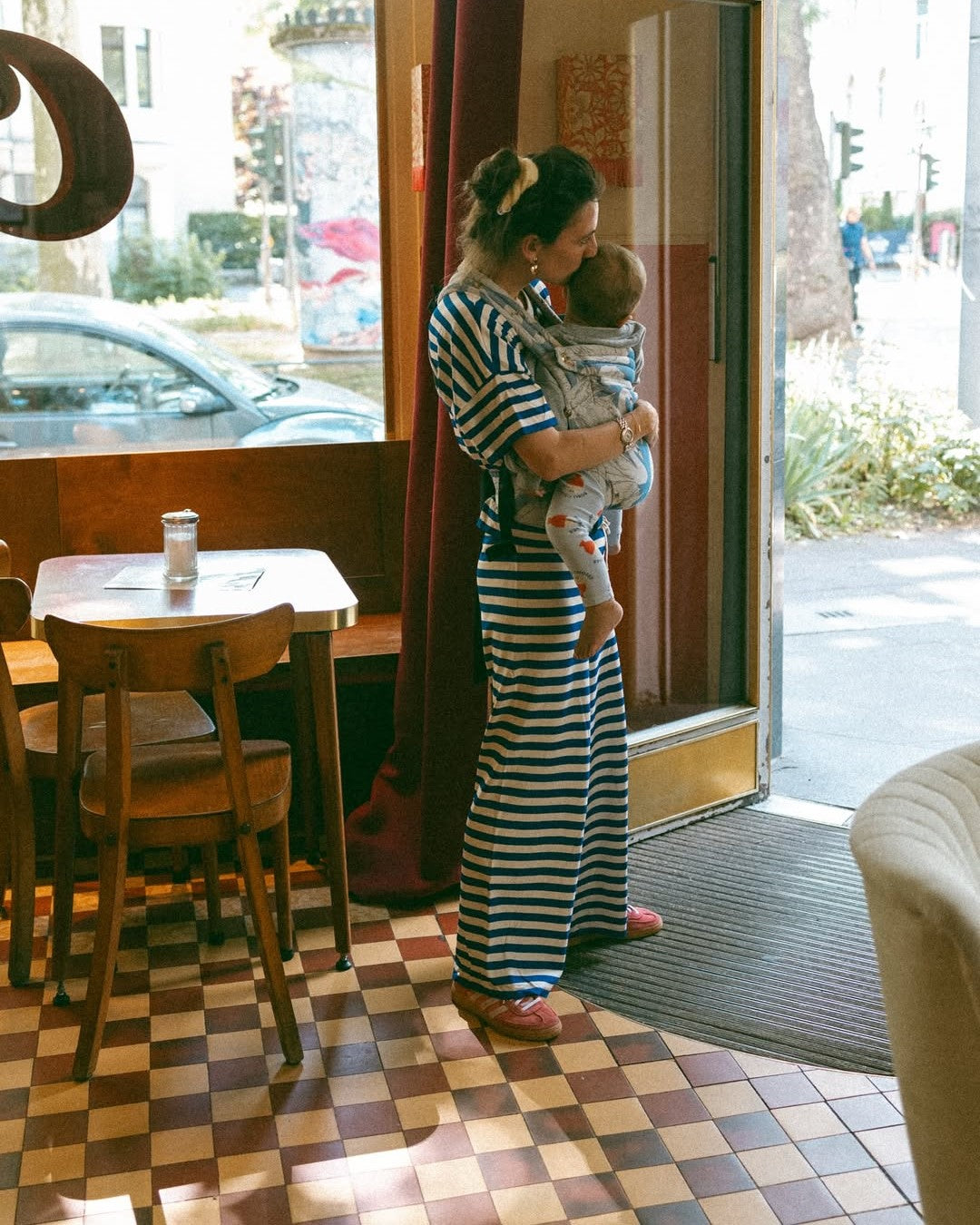 Woman in a striped dress holding a baby in a sling inside a cafe.