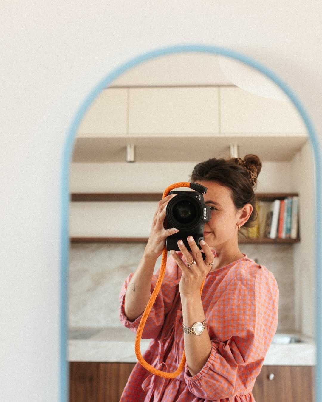 Person taking a mirror selfie with a camera, wearing a pink dress in a kitchen.