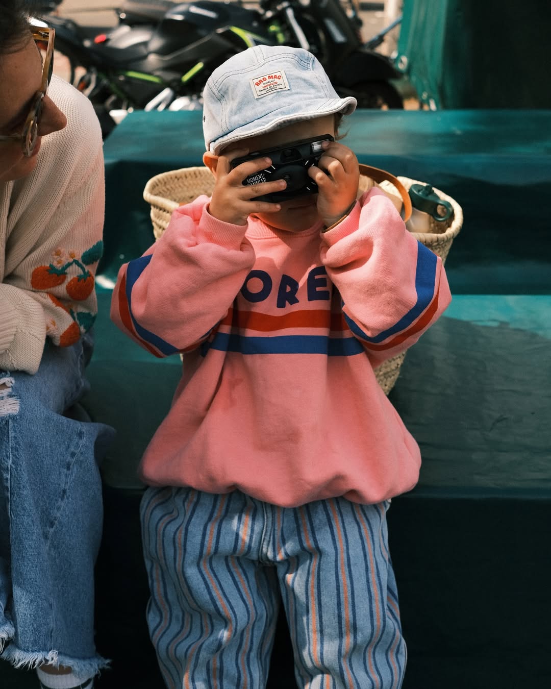 Child in a pink sweatshirt and striped pants holding a phone, with a person partially visible on the left.