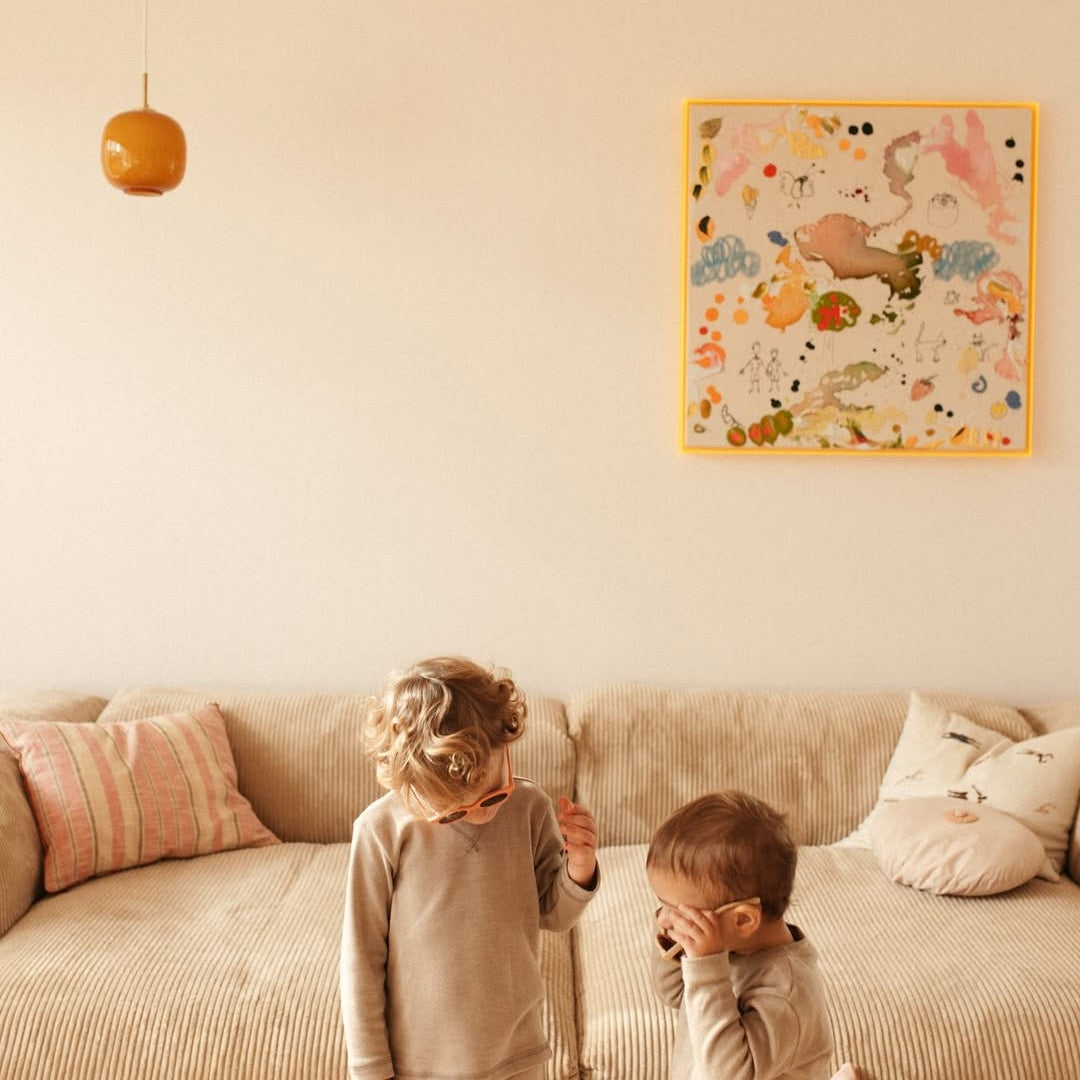 Two children standing on a couch in a room with a colorful wall art piece.