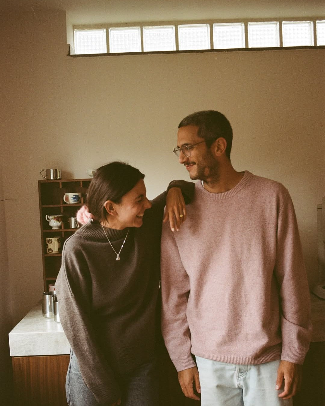 Two people standing close together in a room with a window above them.