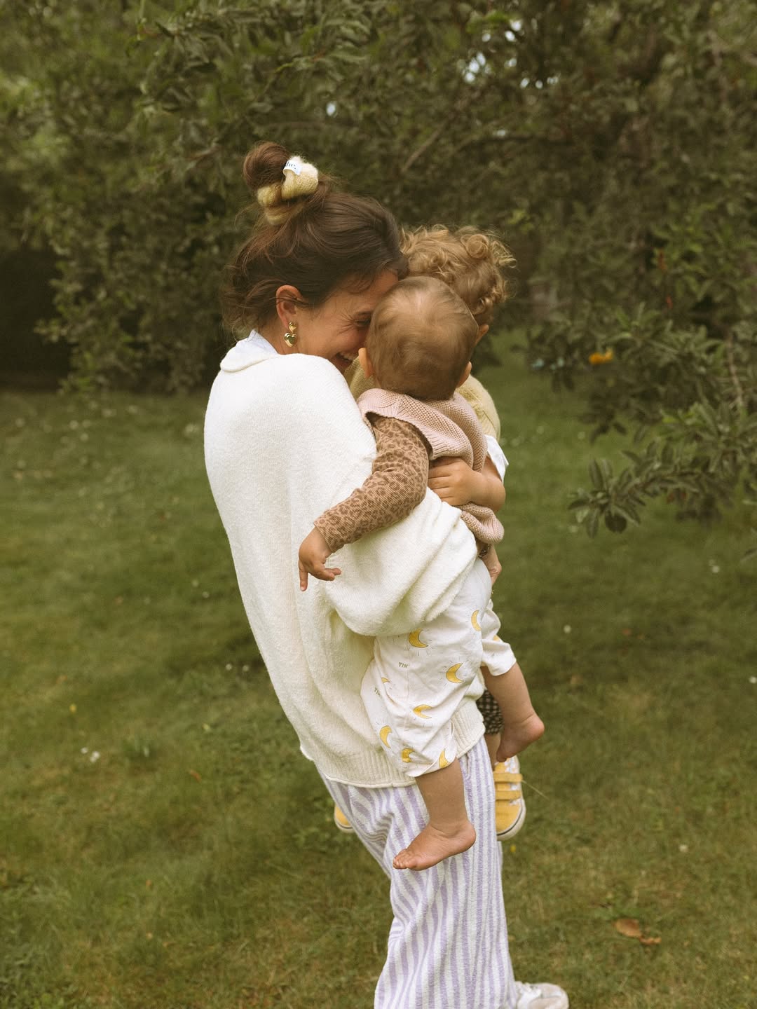 Woman holding two children outdoors with greenery in the background