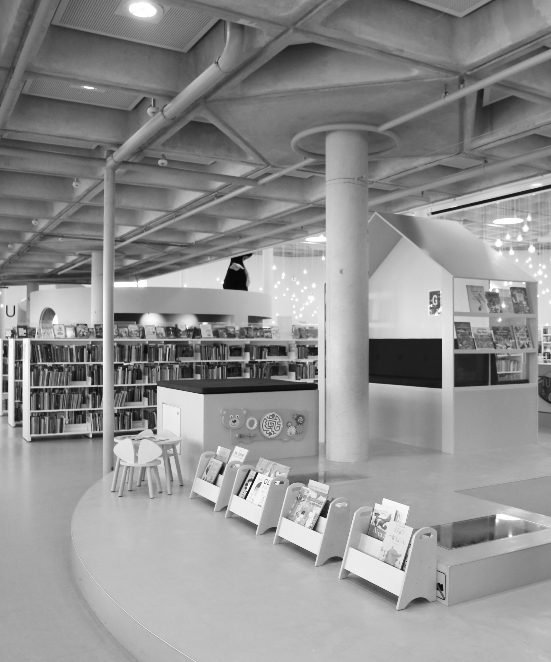 A black and white interior architectural photograph showing a modern library or reading space with bookshelves, reading tables, and a curved floor, with a large white blank space on the right side of the frame.