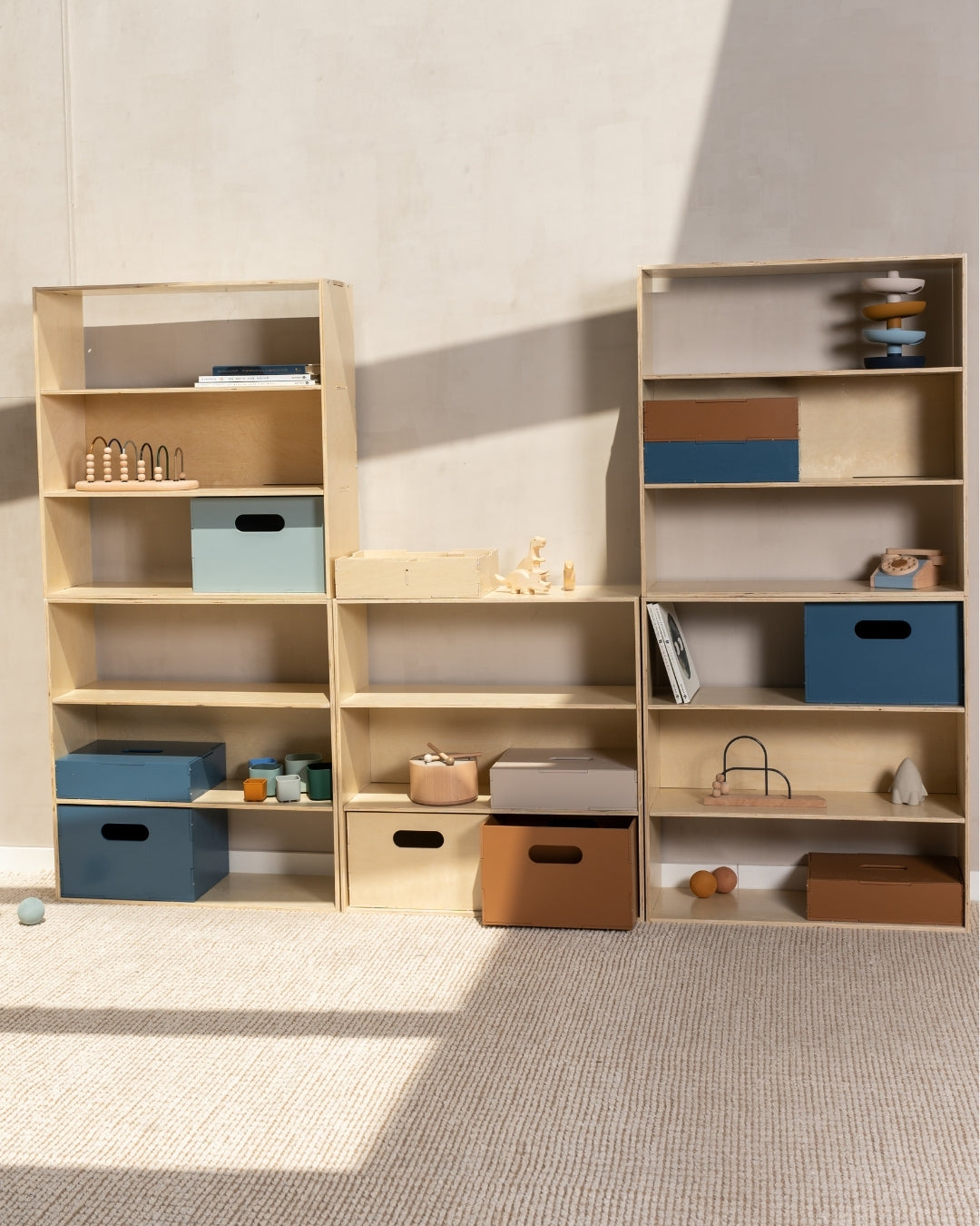 A set of three light natural wood modular bookshelves styled with various colorful storage boxes, toys, and books in a minimalist room with textured beige walls and carpet.