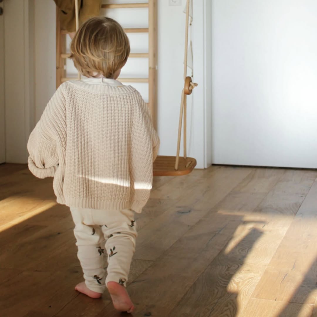 Child in a beige sweater and white pants walking on a wooden floor with a swing in the background.