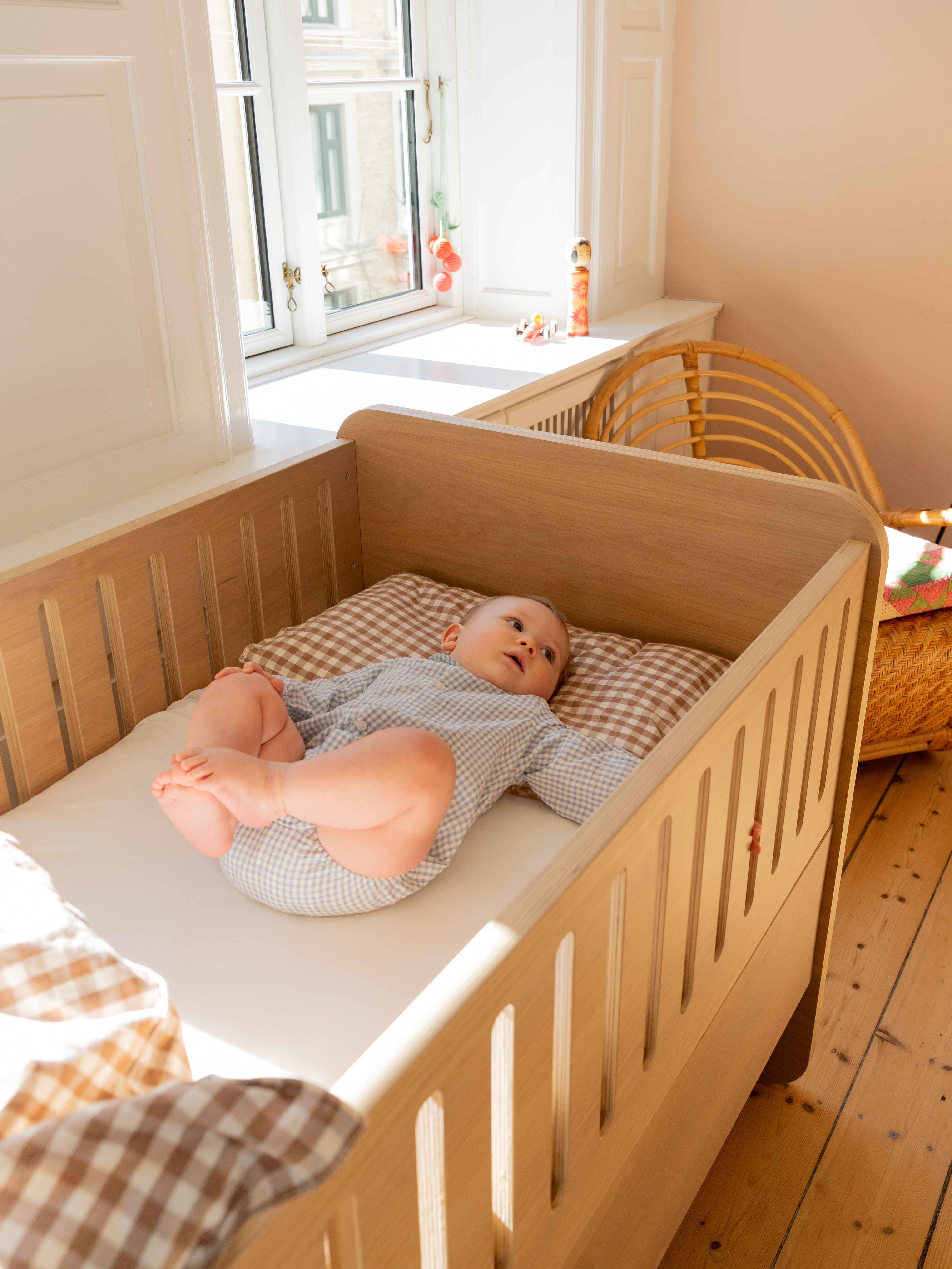 A baby in a light blue and white checkered onesie lies on a mattress inside a light wood crib, with a brown and white checkered pillow. The crib is placed in a warm, sunlit nursery with wooden floors and a rattan chair.