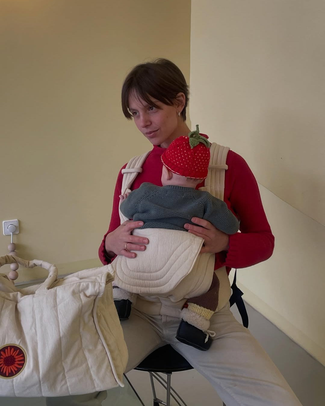 Woman holding a baby in a carrier with a strawberry hat in a room.