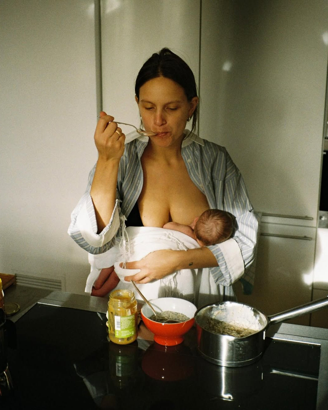 Woman breastfeeding a baby in a kitchen while preparing food.