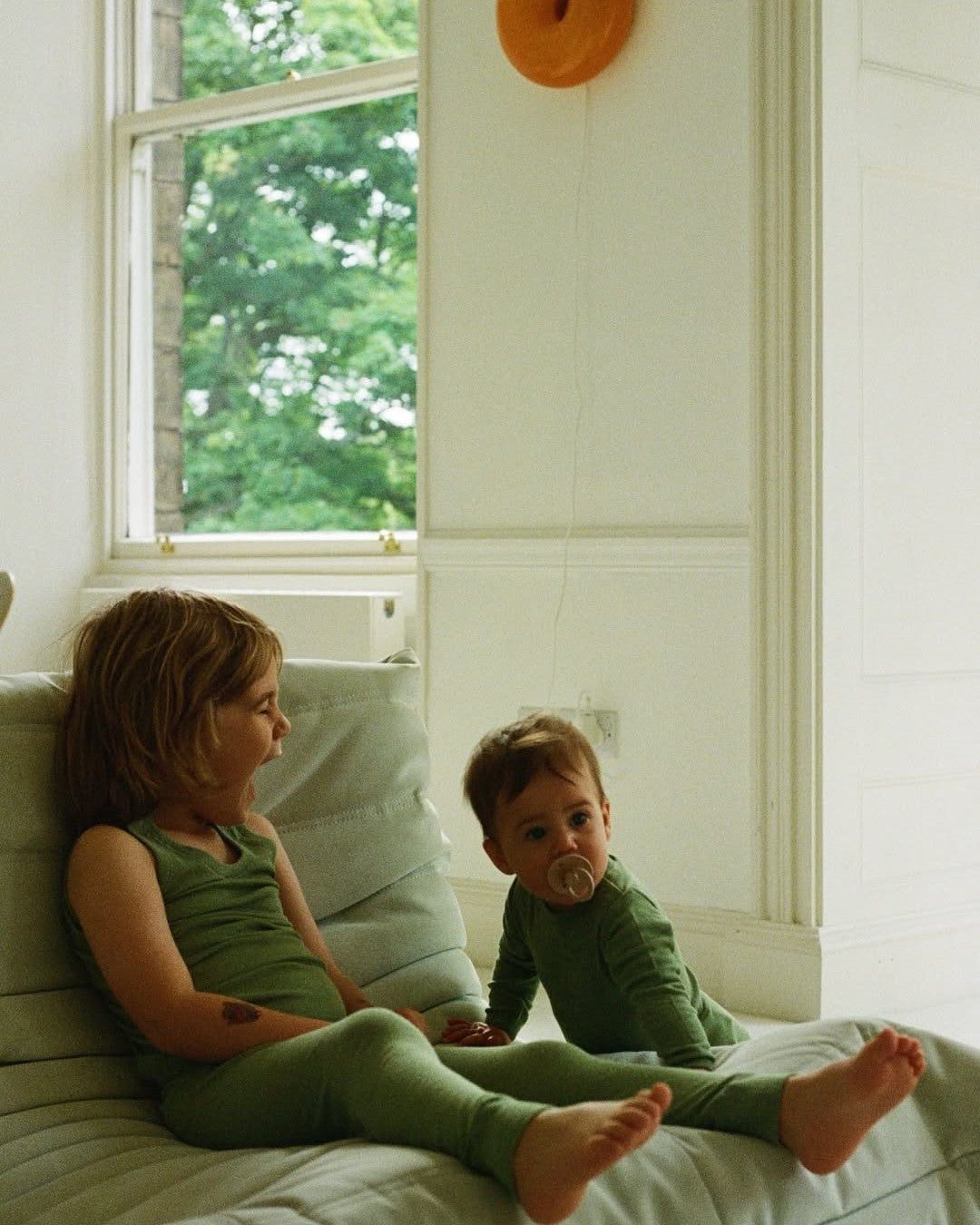 Two children in green outfits sitting on a bed with a window in the background
