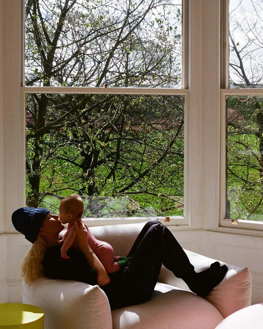 Woman holding a baby on a couch with a large window showing greenery outside