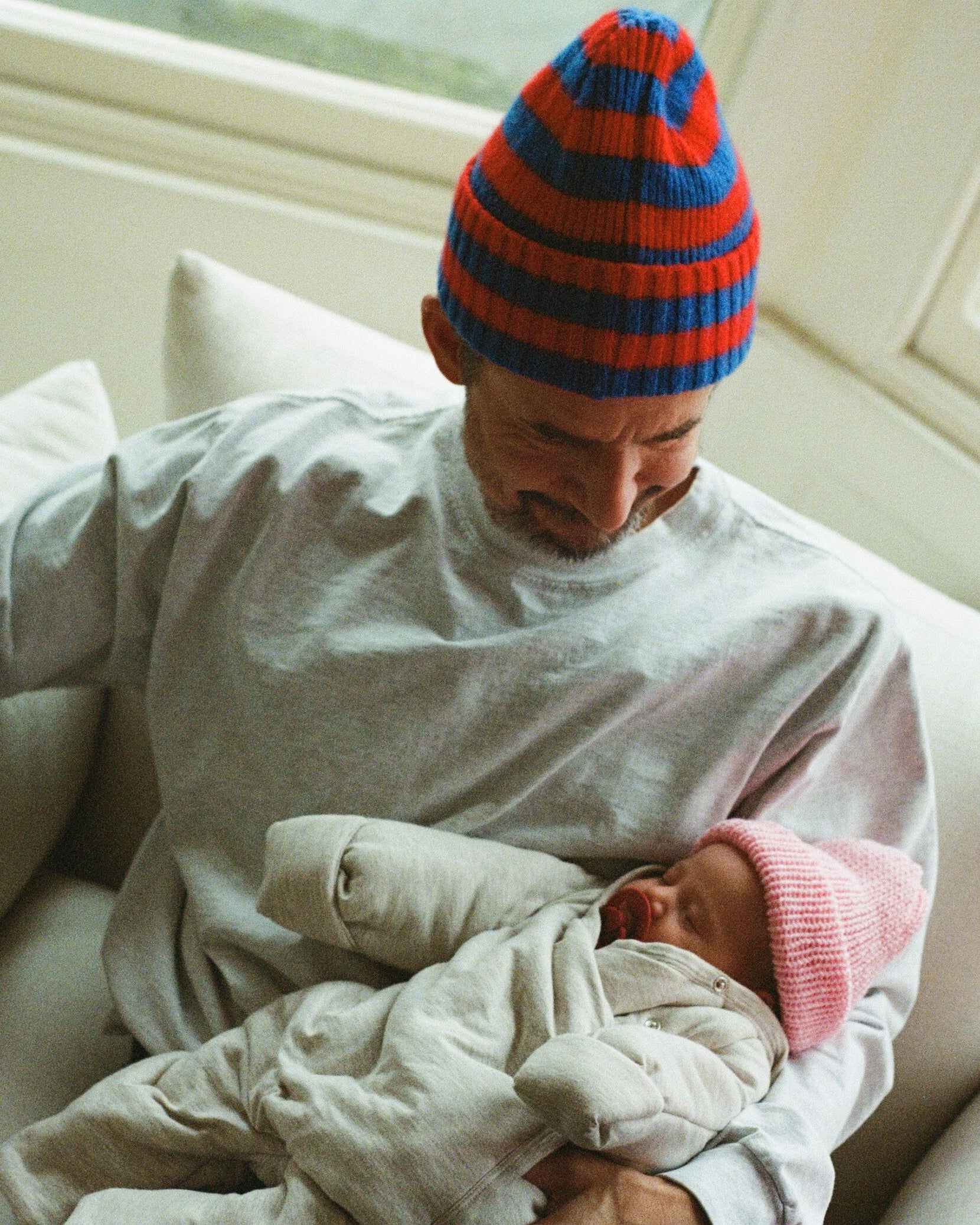 Man and baby sitting on a couch wearing colorful hats in a bright room.