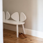 A white children's bench with a curved, shell-like backrest sits on a light wood floor against a neutral wall, with a pair of brown shoes placed underneath.