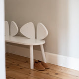 A white children's bench with a curved, shell-like backrest sits on a light wood floor against a neutral wall, with a pair of brown shoes placed underneath.