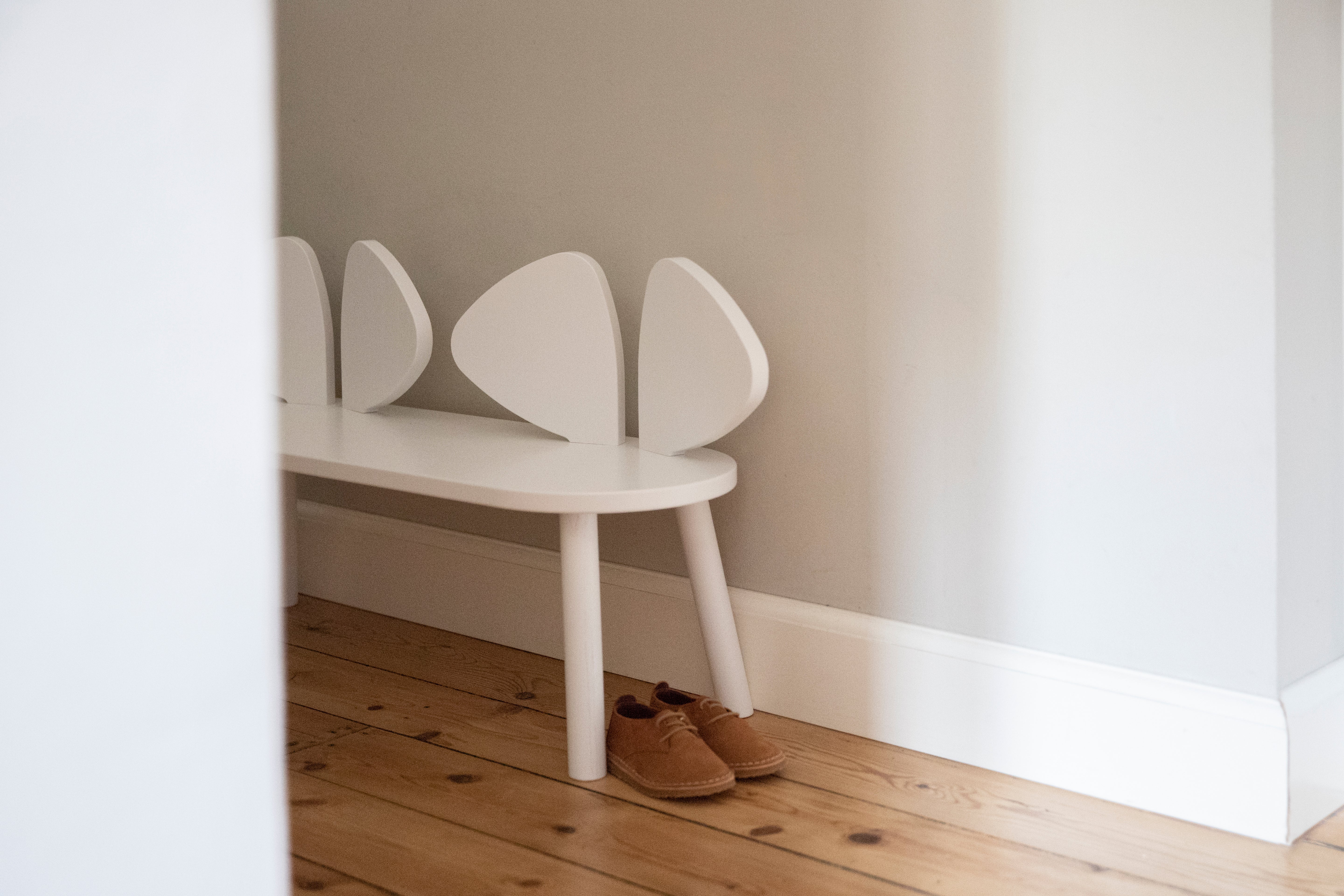 A white children's bench with a curved, shell-like backrest sits on a light wood floor against a neutral wall, with a pair of brown shoes placed underneath.