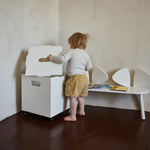 A toddler stands barefoot in a room corner next to a white rolling storage box with a hinged lid, beside a white bench with sculptural shell-shaped backs.