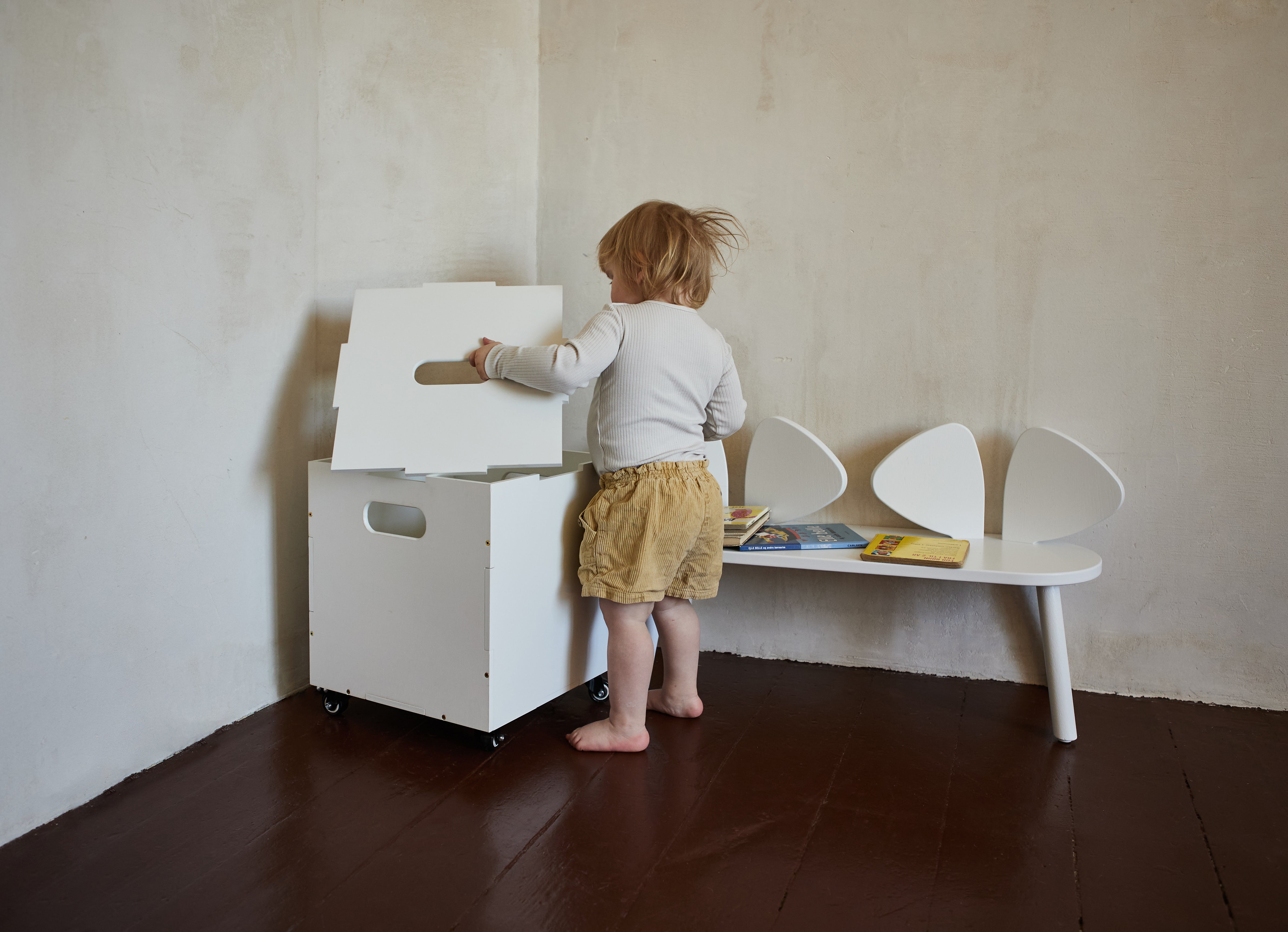 A toddler stands barefoot in a room corner next to a white rolling storage box with a hinged lid, beside a white bench with sculptural shell-shaped backs.