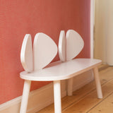 A white wooden bench with a unique leaf-shaped backrest, positioned against a textured coral-pink wall on a light wood floor.