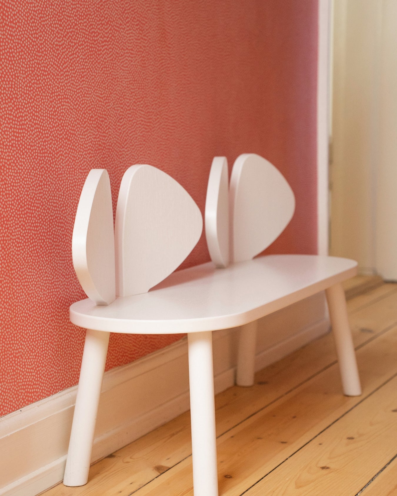 A white wooden bench with a unique leaf-shaped backrest, positioned against a textured coral-pink wall on a light wood floor.