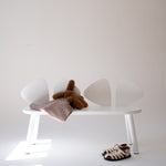 A minimalist white bench with four curved backrests, styled with a brown plush toy, a dusty pink blanket, and a pair of brown-and-white patterned sandals on a white floor against a light beige wall.