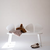 A minimalist white bench with four curved backrests, styled with a brown plush toy, a dusty pink blanket, and a pair of brown-and-white patterned sandals on a white floor against a light beige wall.