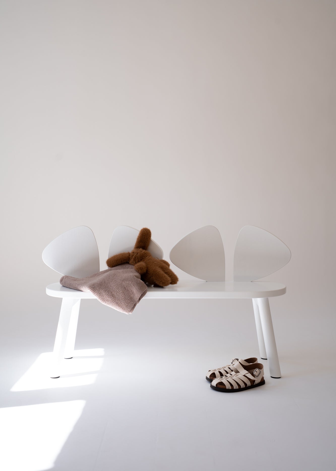 A minimalist white bench with four curved backrests, styled with a brown plush toy, a dusty pink blanket, and a pair of brown-and-white patterned sandals on a white floor against a light beige wall.
