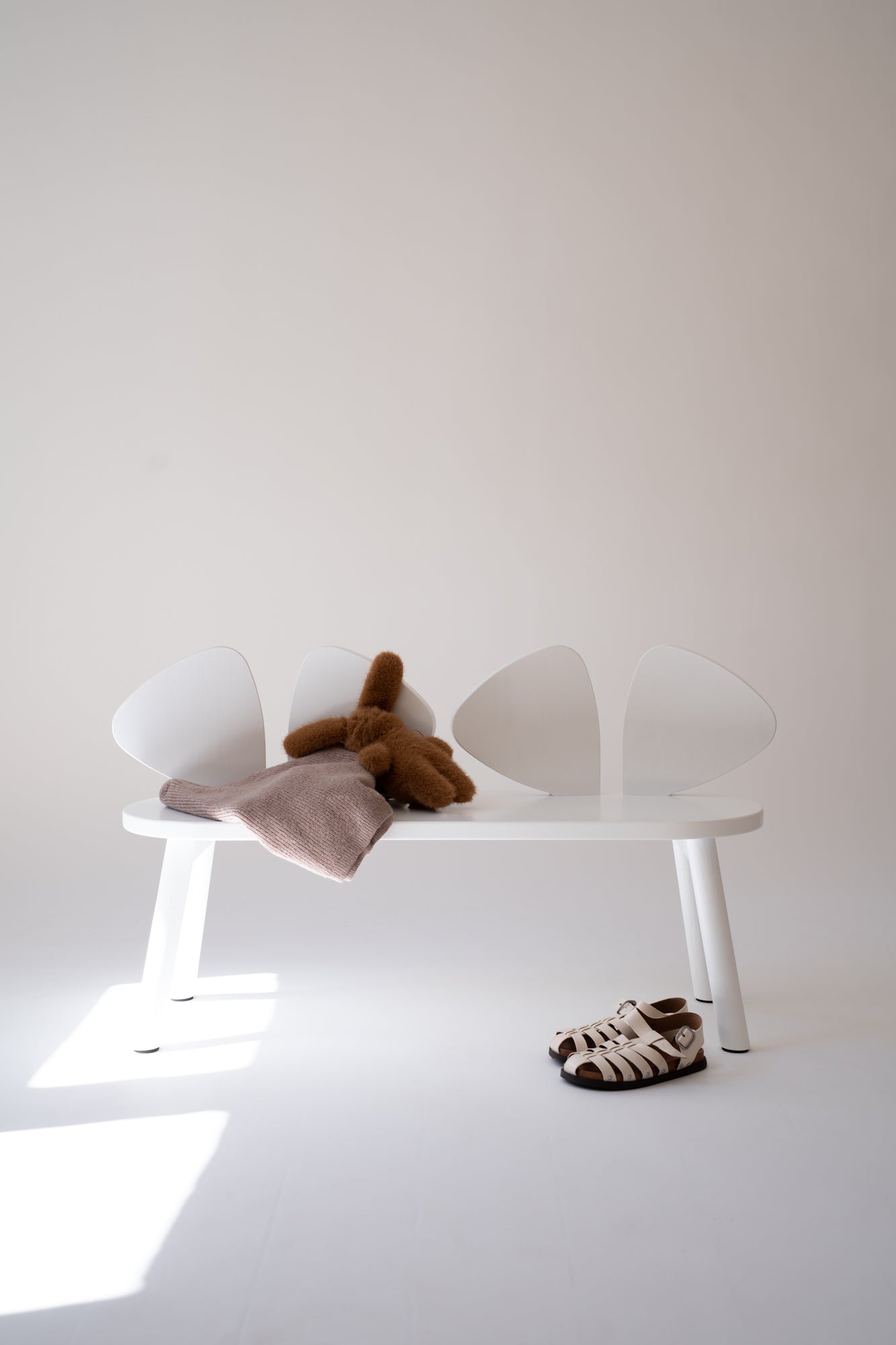 A minimalist white bench with four curved backrests, styled with a brown plush toy, a dusty pink blanket, and a pair of brown-and-white patterned sandals on a white floor against a light beige wall.