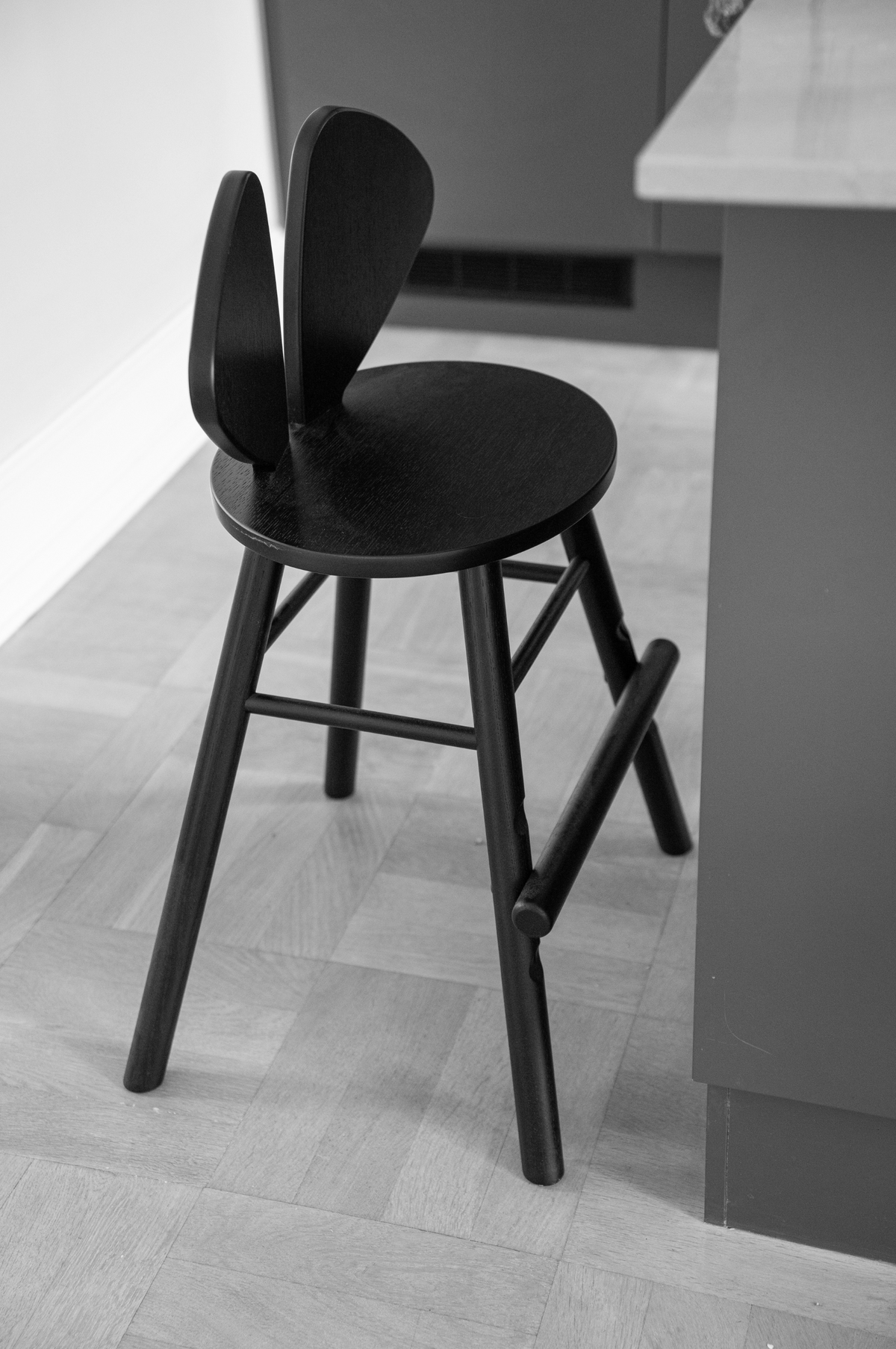 A black wooden bar stool with a unique wingback-style backrest, shown in a modern kitchen setting with a light wood floor and gray cabinetry.