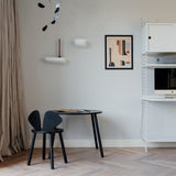 A modern interior room scene with a black round table and black chair near a beige curtain, plus a white shelving unit with a computer monitor and framed wall art.