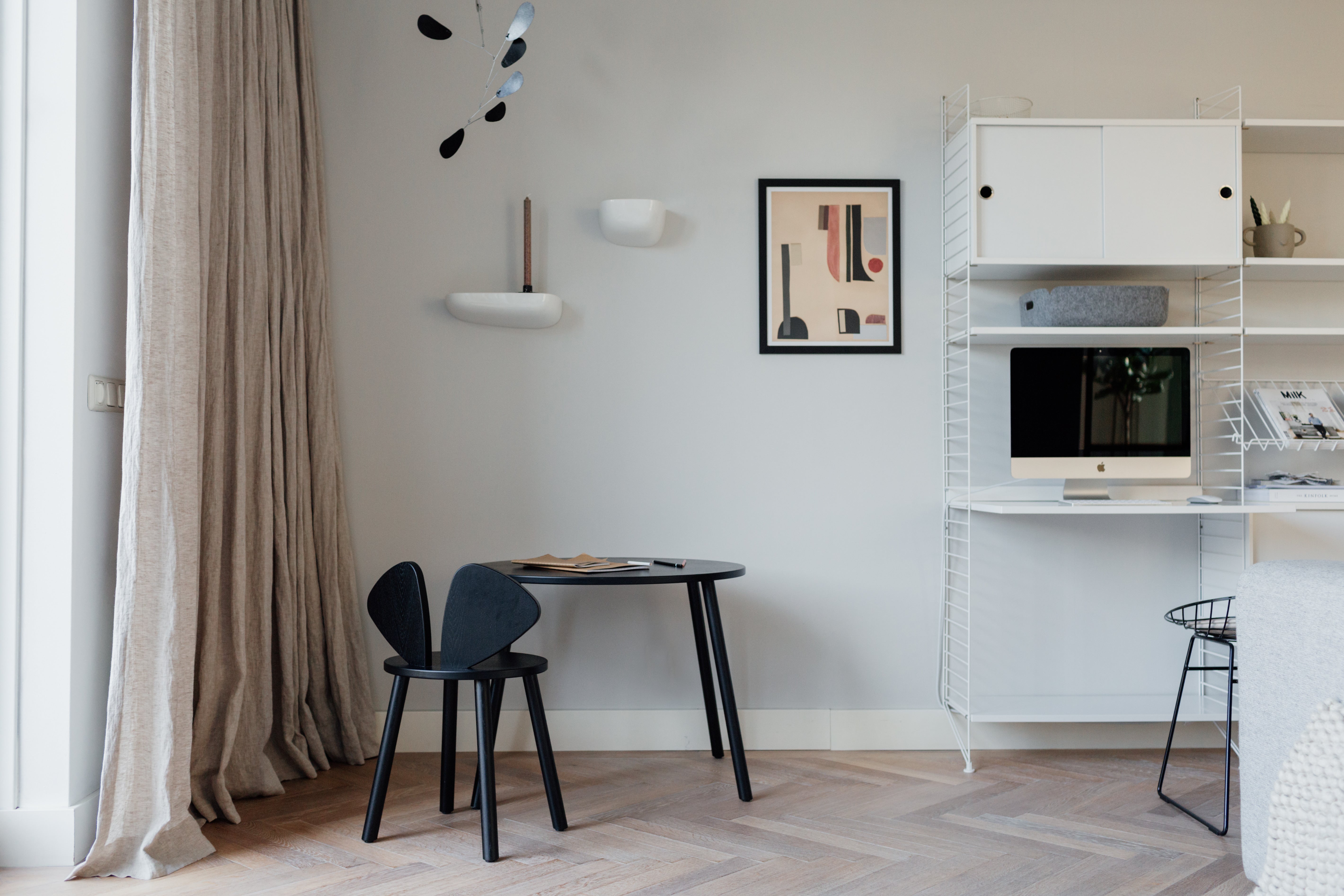 A modern interior room scene with a black round table and black chair near a beige curtain, plus a white shelving unit with a computer monitor and framed wall art.