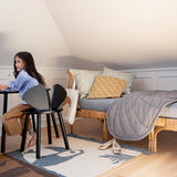 A young girl sits at a small black desk on a chair in a cozy, sunlit attic bedroom. The room features a wooden daybed with gray bedding, patterned pillows, and a rug on a light wood floor.