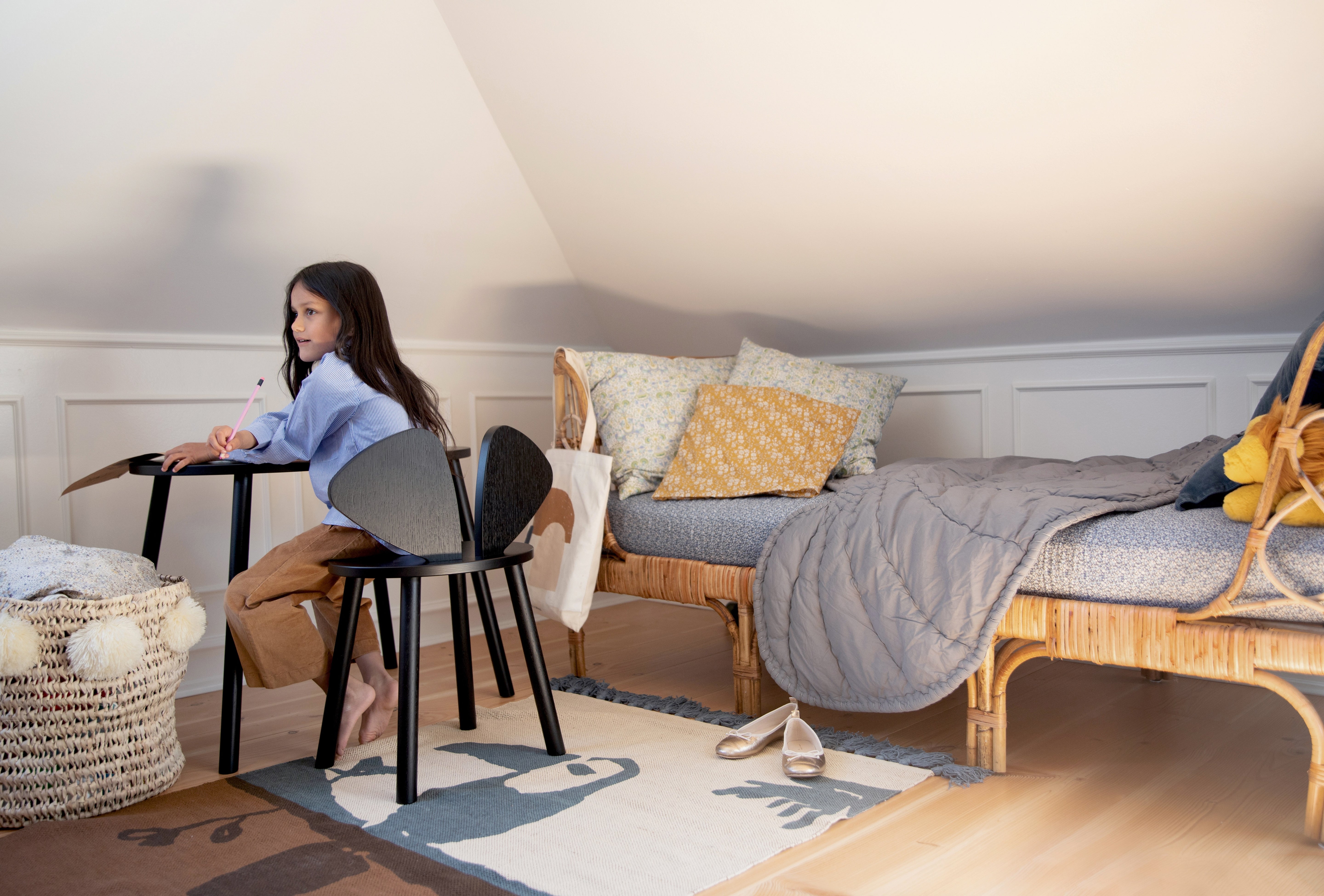A young girl sits at a small black desk on a chair in a cozy, sunlit attic bedroom. The room features a wooden daybed with gray bedding, patterned pillows, and a rug on a light wood floor.