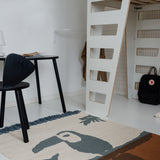 A modern room scene featuring a black molded plastic chair with a unique wing-like back, a black desk, a white loft bed frame, and a beige rug with a dark blue and brown toucan print.