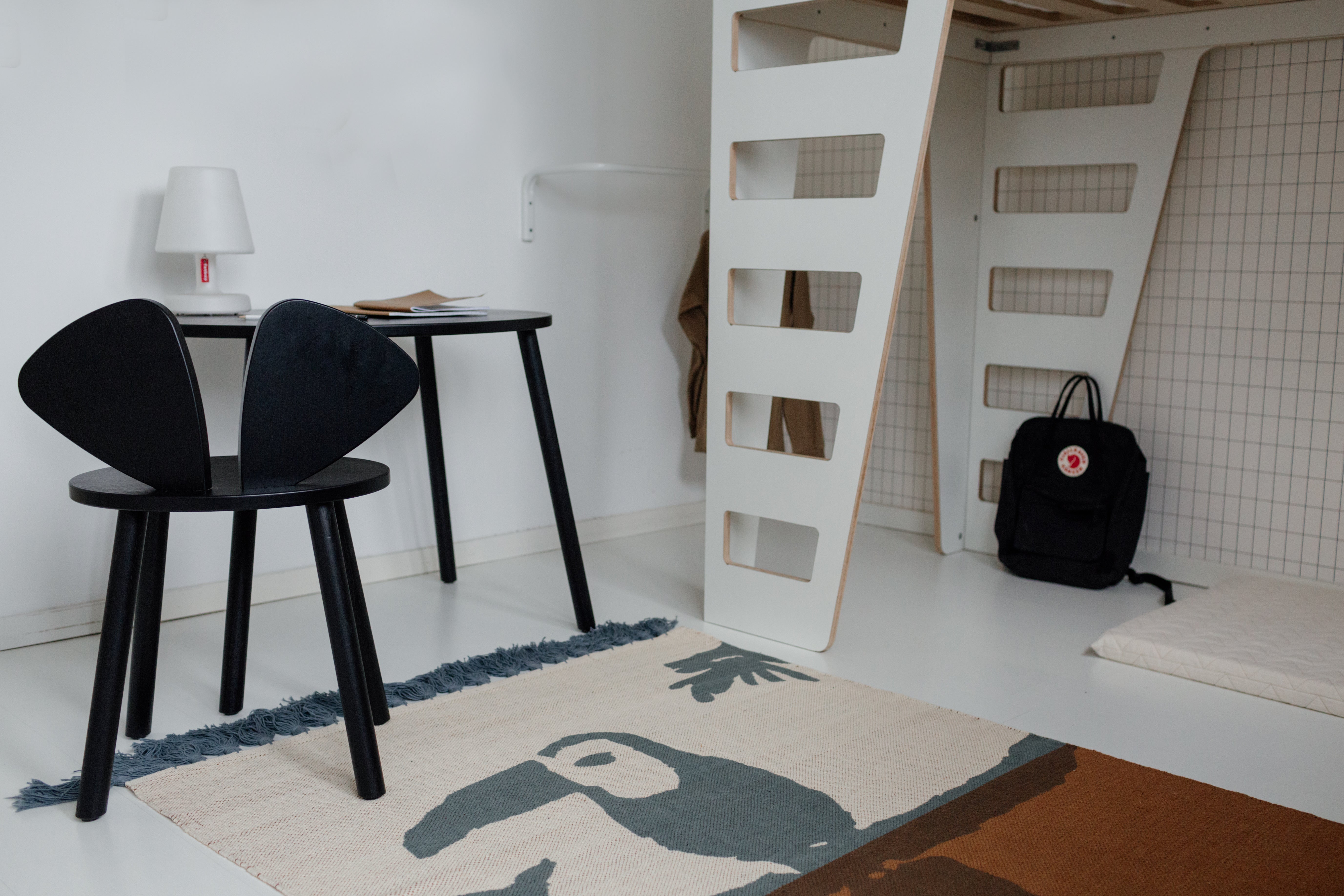 A modern room scene featuring a black molded plastic chair with a unique wing-like back, a black desk, a white loft bed frame, and a beige rug with a dark blue and brown toucan print.