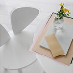 A minimalist white table and chair setup in a bright interior, with a small clear glass vase holding yellow flowers and a stack of paper items on the table.