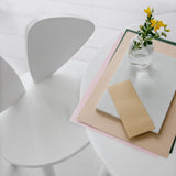 A minimalist white table and chair setup in a bright interior, with a small clear glass vase holding yellow flowers and a stack of paper items on the table.