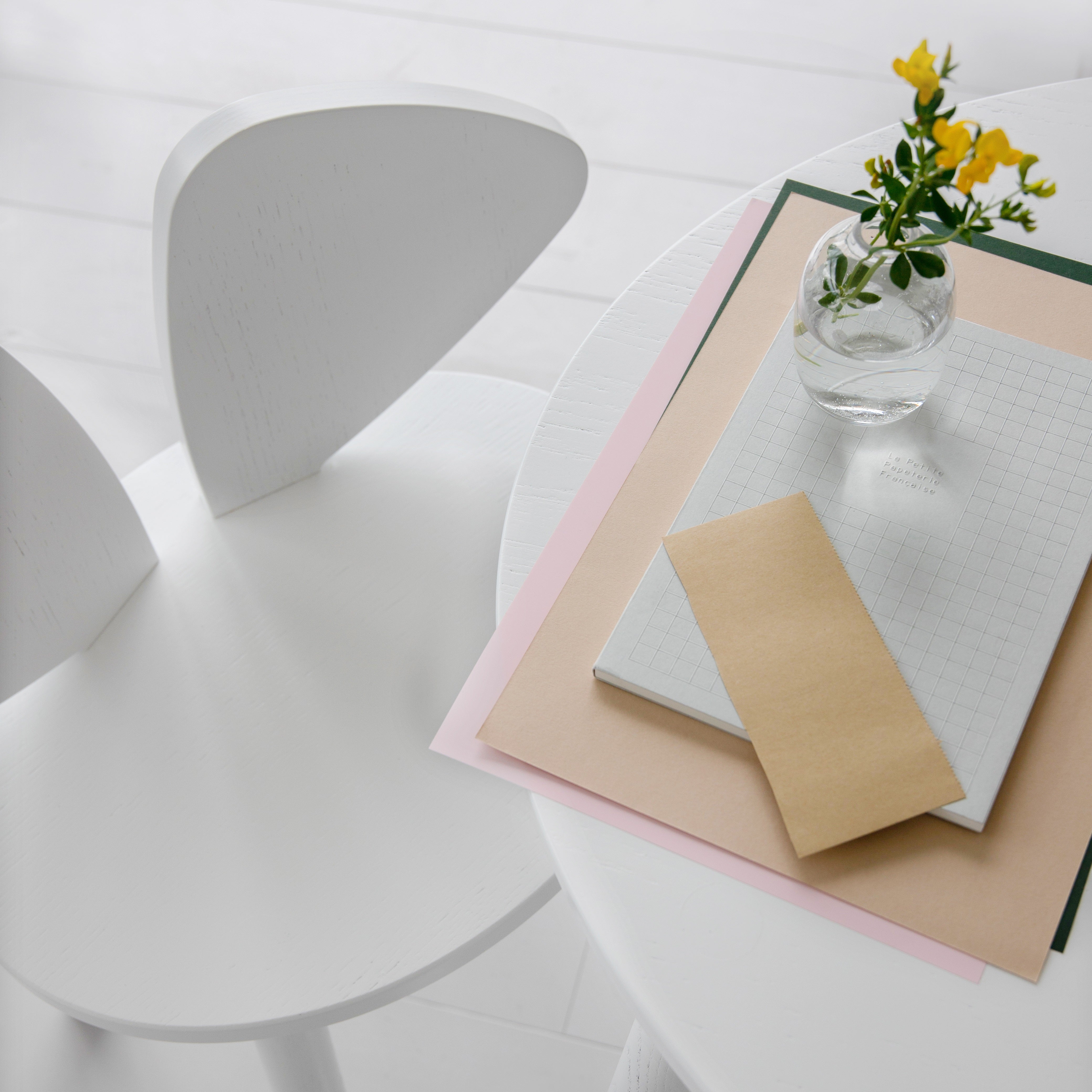 A minimalist white table and chair setup in a bright interior, with a small clear glass vase holding yellow flowers and a stack of paper items on the table.