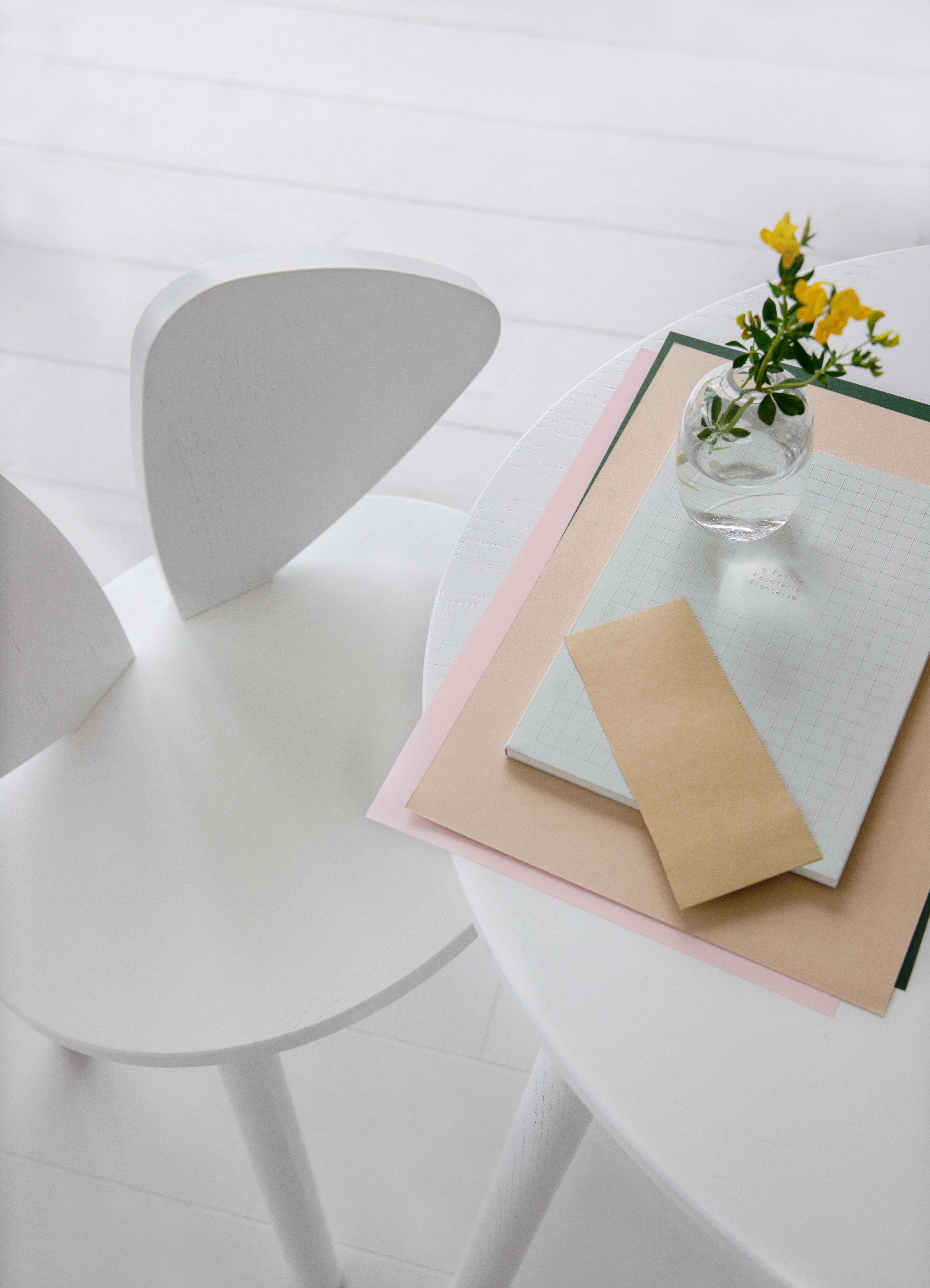 A minimalist white table and chair setup in a bright interior, with a small clear glass vase holding yellow flowers and a stack of paper items on the table.