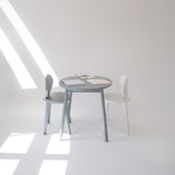 A minimalist children's table set featuring a round gray table and two chairs, one gray and one white, in a bright, sunlit room with geometric shadows.