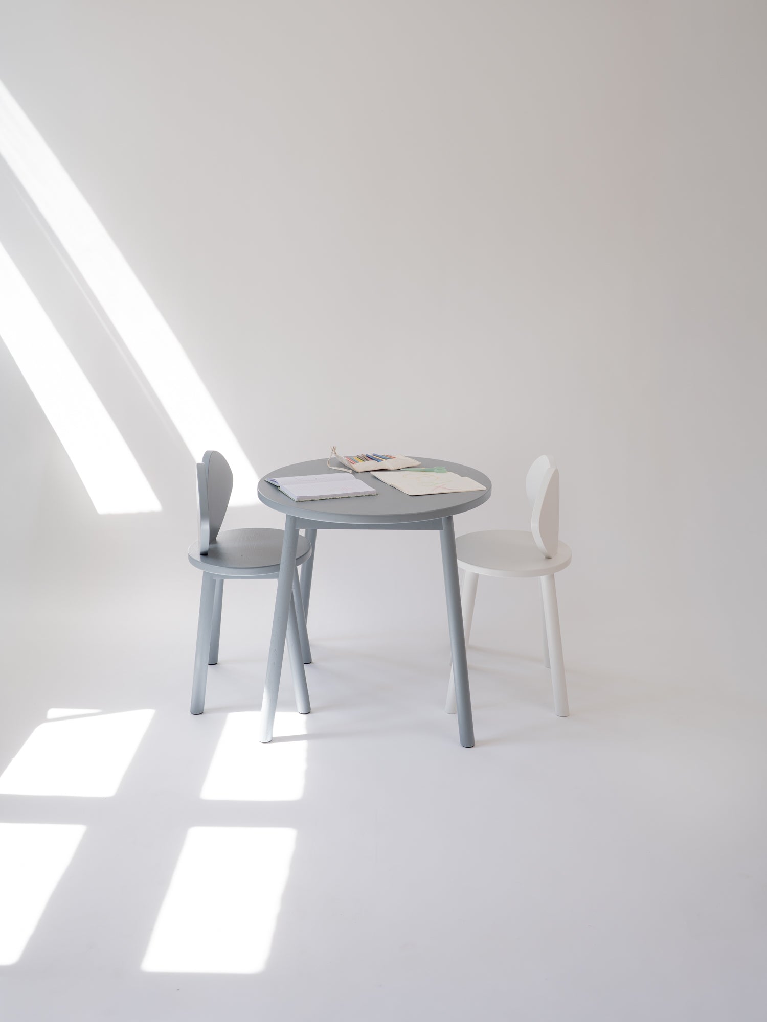 A minimalist children's table set featuring a round gray table and two chairs, one gray and one white, in a bright, sunlit room with geometric shadows.