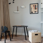 A styled interior corner featuring a black round table, a black chair, a light wood rolling storage box on wheels, beige curtains, and a framed abstract print on a light gray wall.