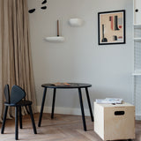 A styled interior corner featuring a black round table, a black chair, a light wood rolling storage box on wheels, beige curtains, and a framed abstract print on a light gray wall.