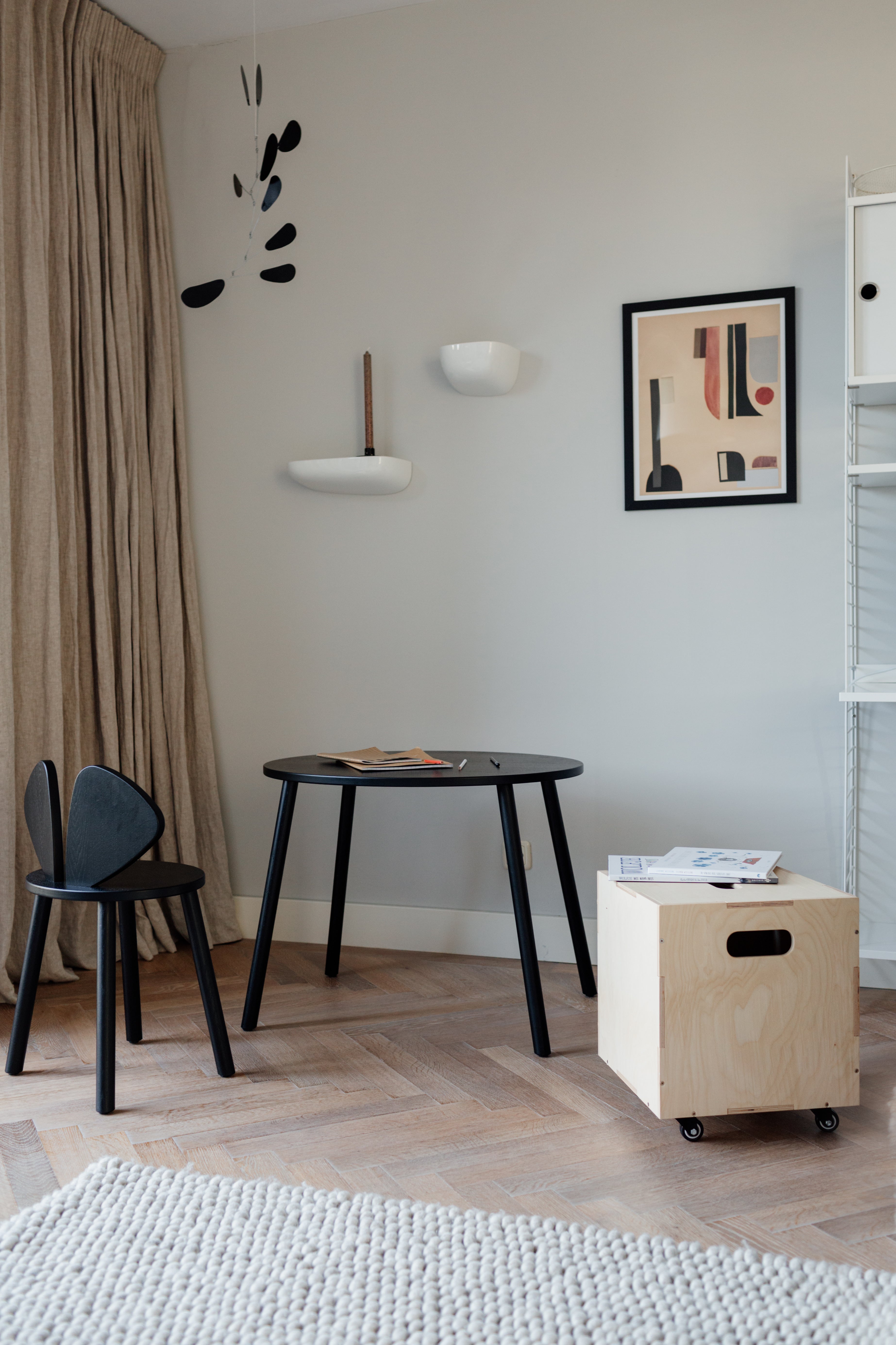 A styled interior corner featuring a black round table, a black chair, a light wood rolling storage box on wheels, beige curtains, and a framed abstract print on a light gray wall.