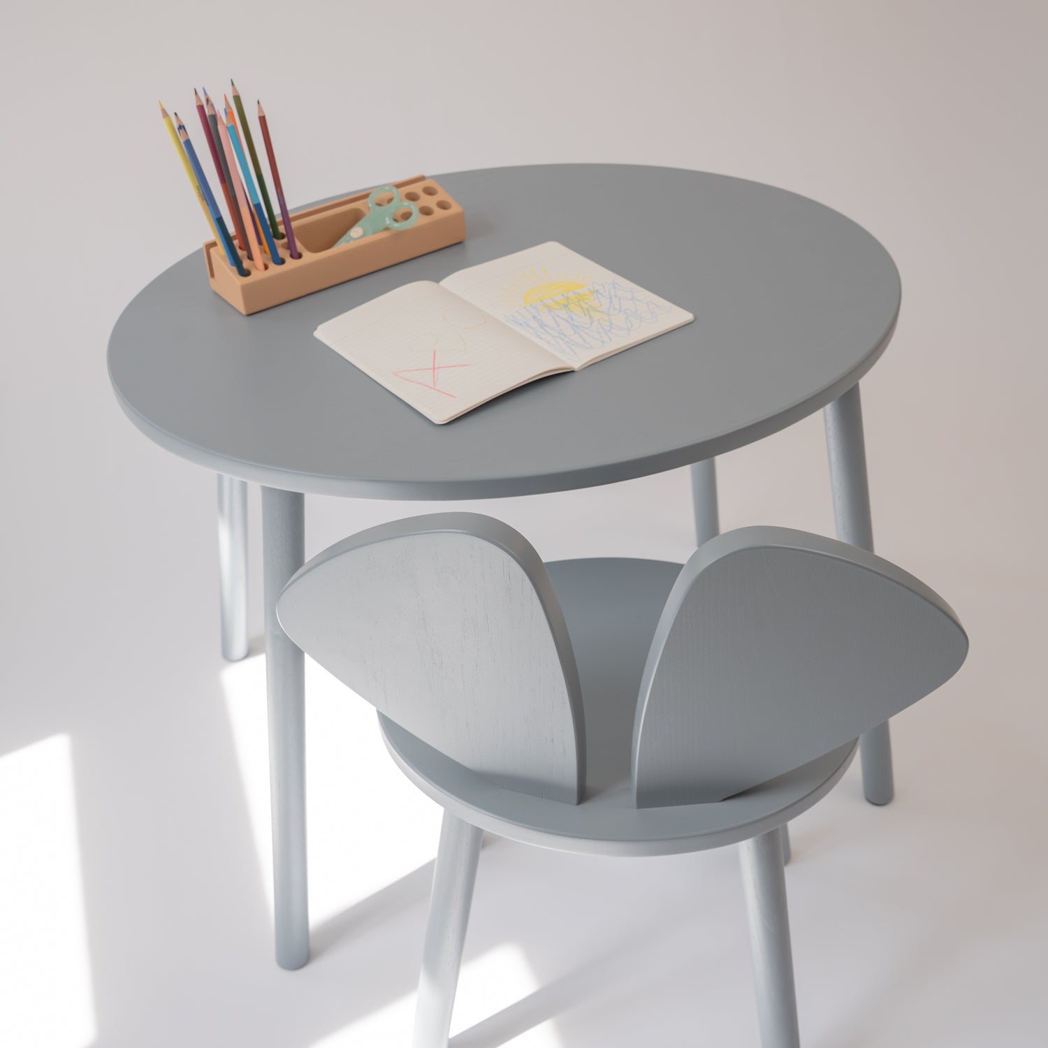 A minimalist children's study set featuring a round gray table and a matching chair with wing-like backrests, styled with a pencil holder and an open notebook on a clean white background.