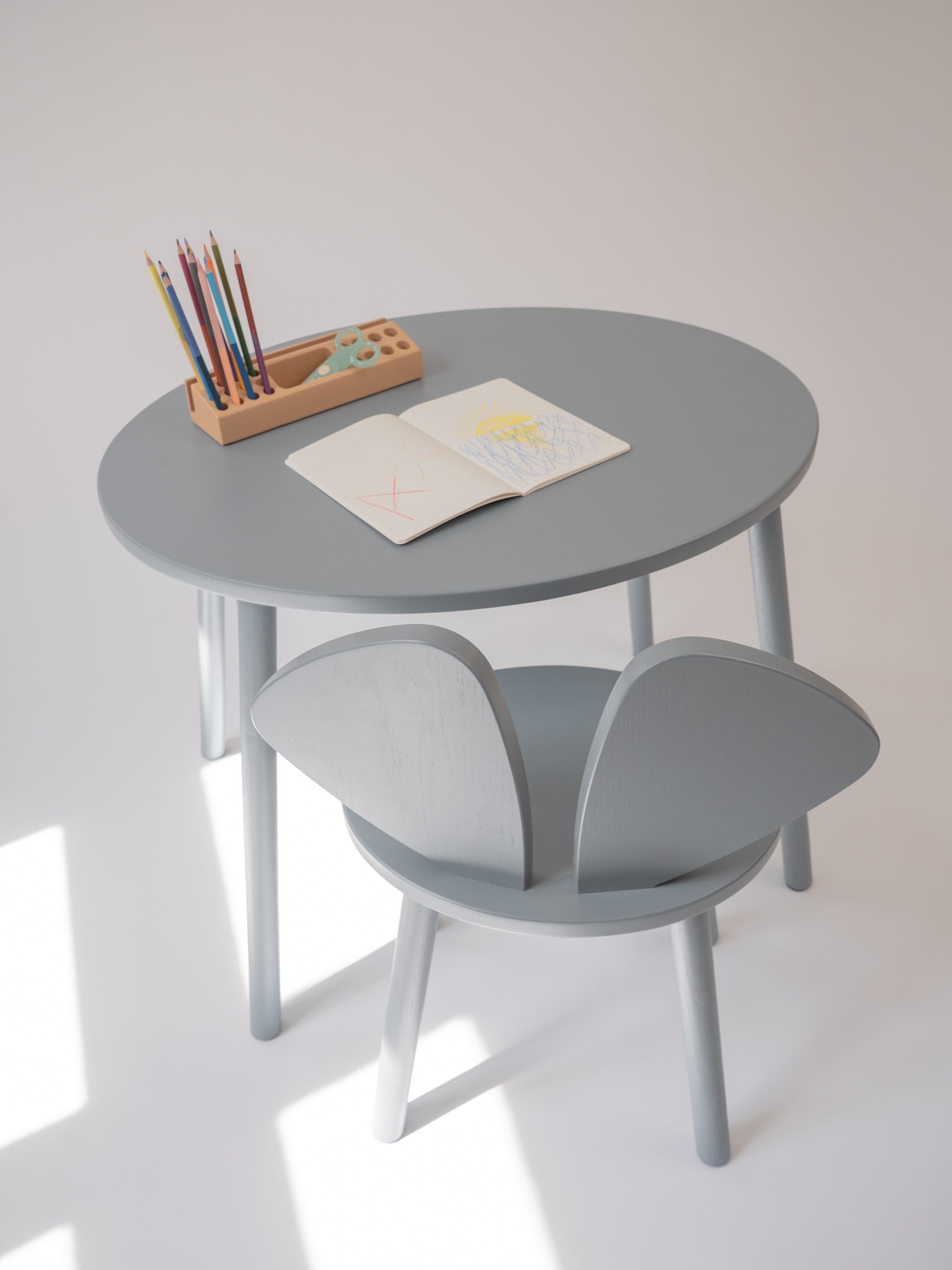 A minimalist children's study set featuring a round gray table and a matching chair with wing-like backrests, styled with a pencil holder and an open notebook on a clean white background.