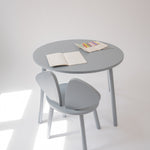 A minimalist light gray children's table and chair set with open books on the tabletop, photographed in a bright white room with natural window light casting soft shadows.