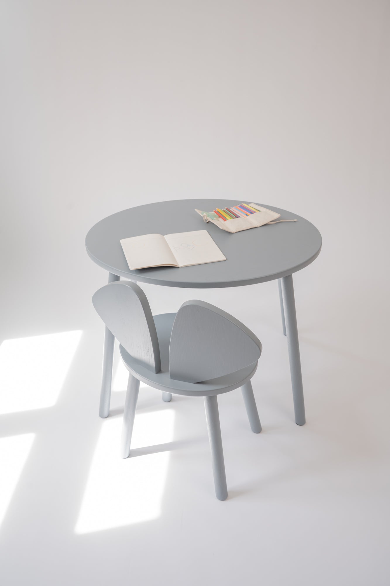 A minimalist light gray children's table and chair set with open books on the tabletop, photographed in a bright white room with natural window light casting soft shadows.