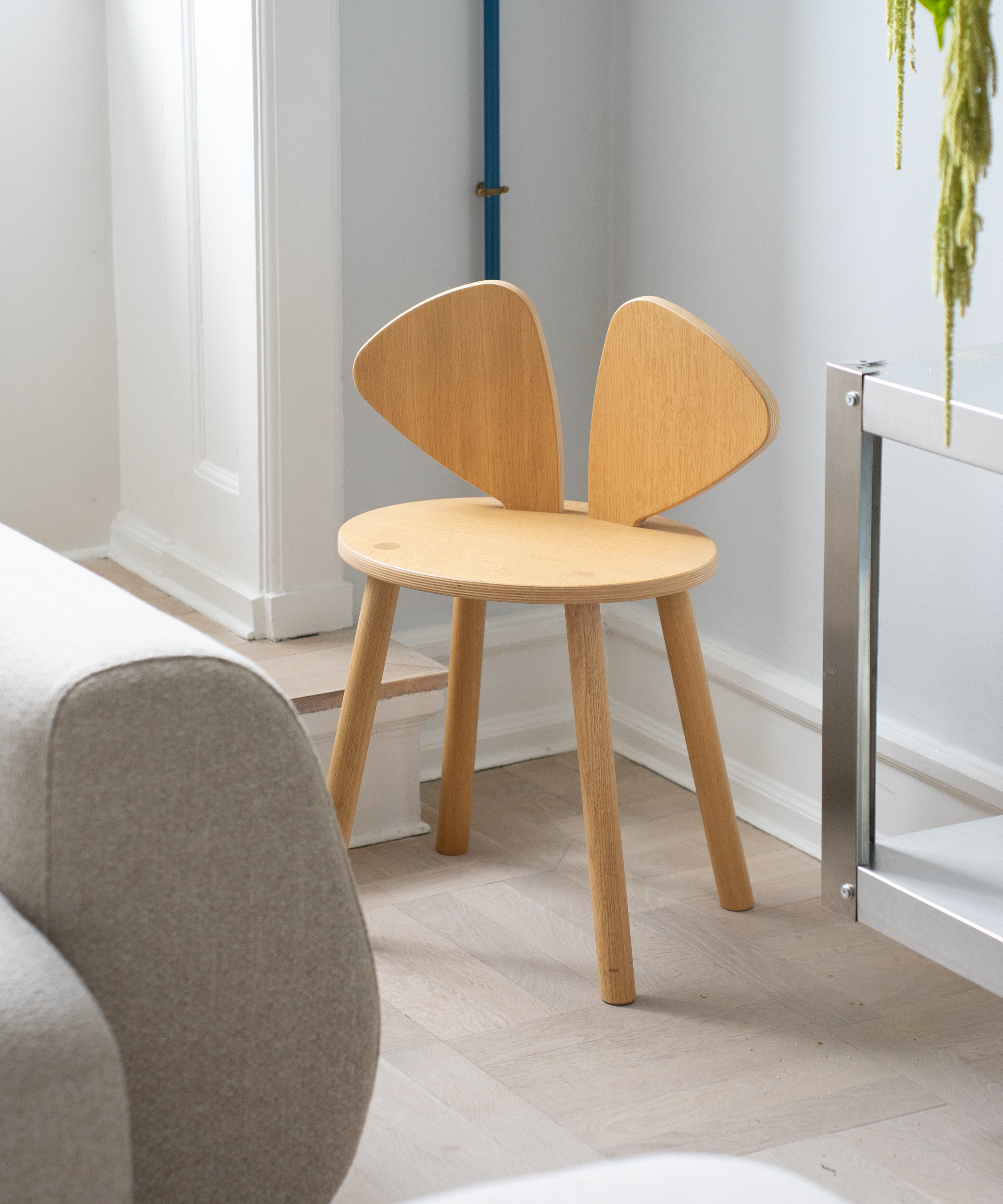 A light natural wood children's stool with a round seat and two curved backrest wings, shown in a bright, modern room next to a gray sofa.