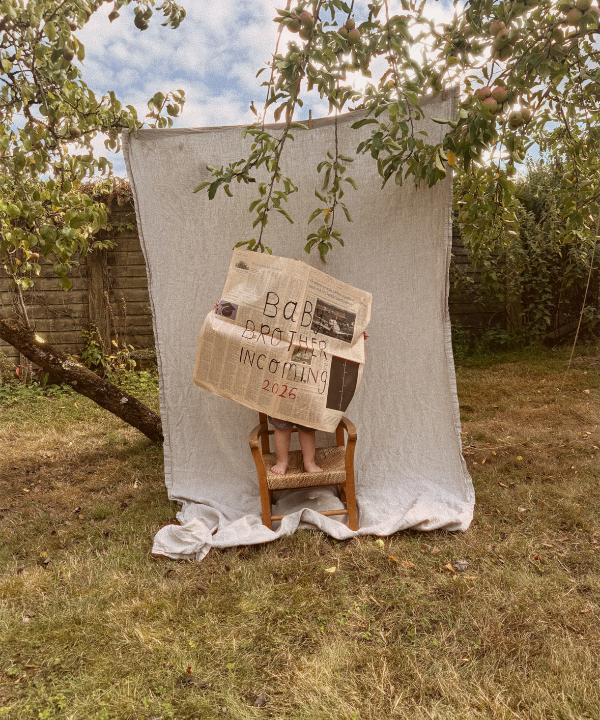 Person sitting on a chair holding a newspaper with text, standing behind a white curtain in an outdoor setting.